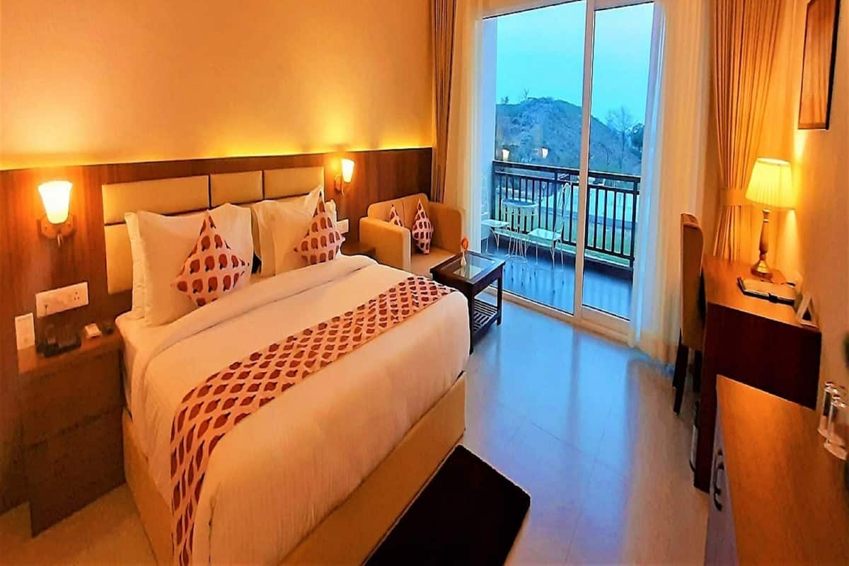 Habitación de hotel con una cama grande, una mesita, una silla, un balcón con vistas y una mesita de noche con lámpara.