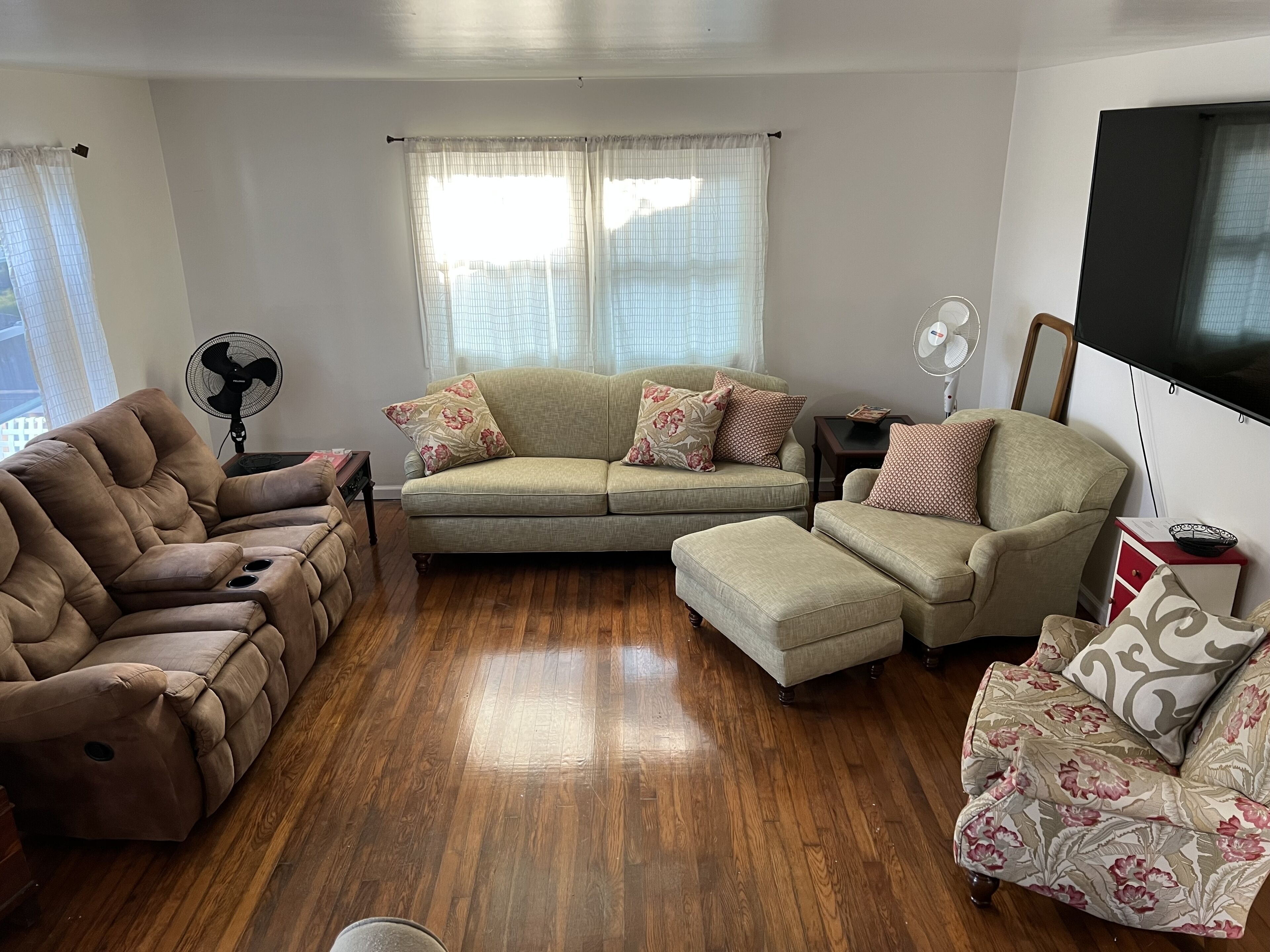 A living room with a brown leather sofa, a beige sectional sofa, and a patterned armchair. There is a wooden floor, a ceiling fan, and a flat-screen TV mounted on the wall.