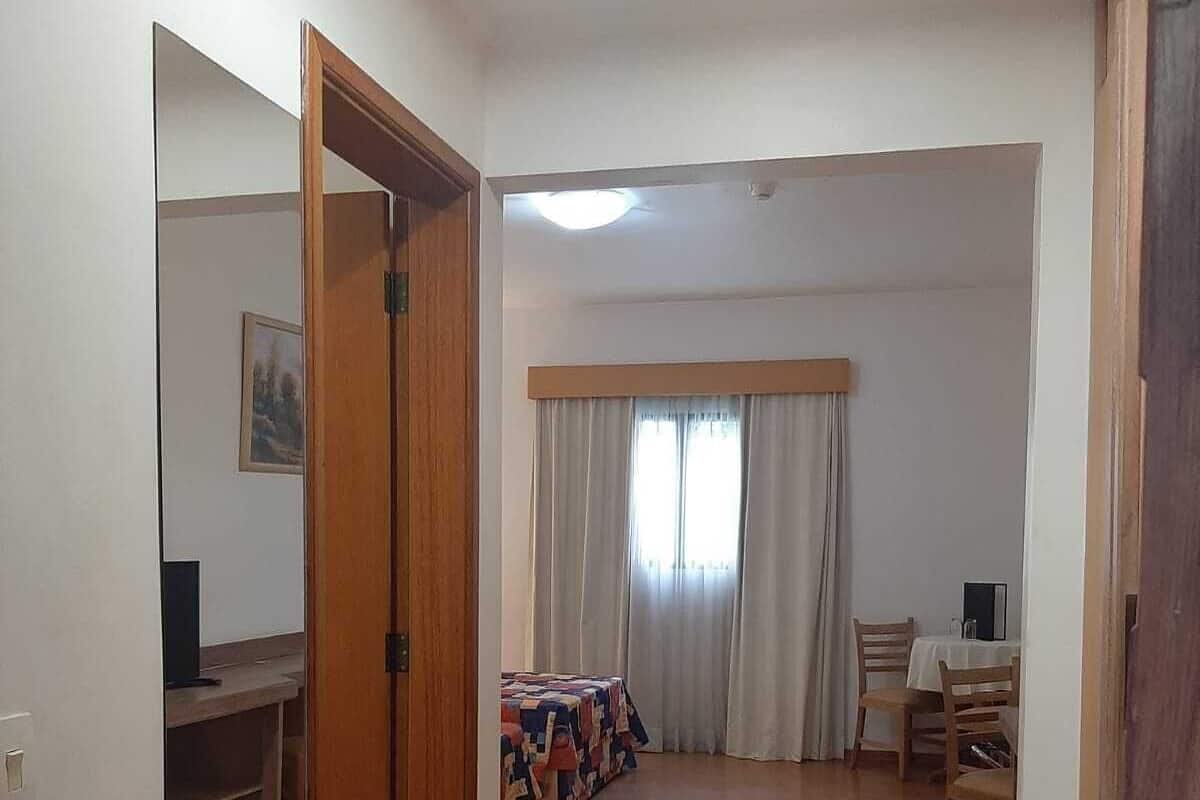 Una habitación con piso de madera, una cama con ropa de cama estampada, un escritorio con silla, una ventana con cortinas y un espejo en la pared.