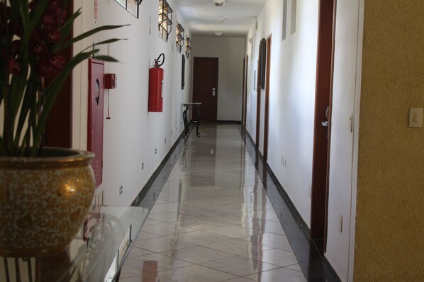 A hallway with a polished tile floor, a potted plant, a fire extinguisher, and a door at the end. - PARADIES HOTEL EXPRESS (Jarinu)