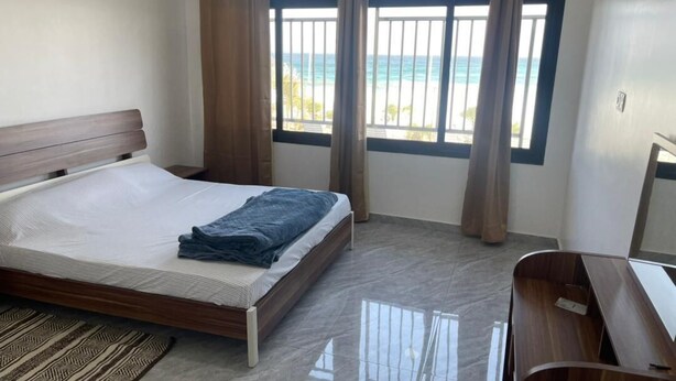 A bedroom with a bed, a desk, and a view of the beach through large windows. - SEA VIEW APARTMENT (DAKAR)