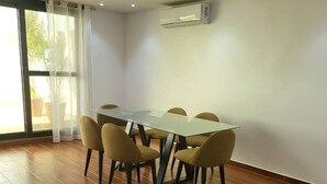 A dining area with a rectangular table and six chairs, a large window with curtains, and an air conditioning unit on the wall. - SEA VIEW APARTMENT (DAKAR)