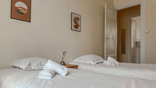 Een hotelkamer met twee bedden, een nachtkastje met een lamp, en een deur naar een andere kamer.