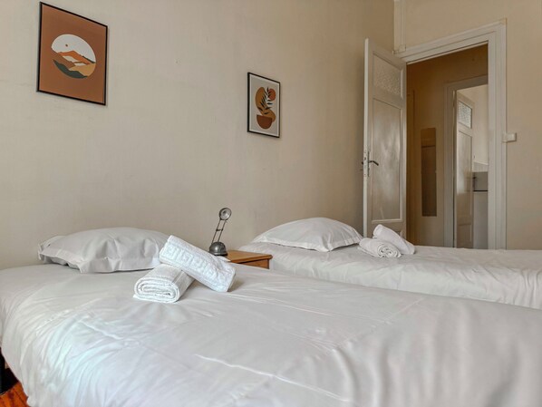 A hotel room with two beds, a bedside table with a lamp, and a door leading to another room.