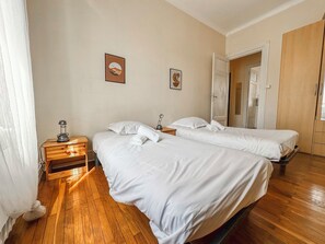 A hotel room with two beds, wooden flooring, a nightstand with a lamp, and a door leading to another room.
