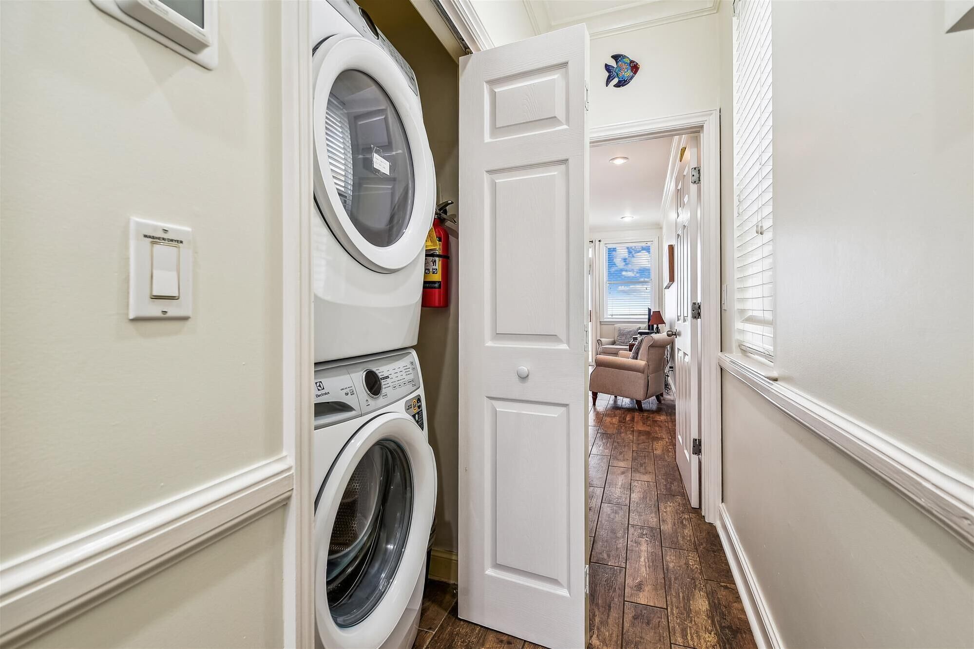 A laundry room with stacked washers and dryers, a fire extinguisher, and a door leading to a living area with a couch and a view of the sky.