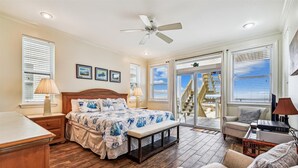A bedroom with a bed, bedside tables, a ceiling fan, and a view of the beach through large windows. - Welcome to The Sandbox! (St. George Island)