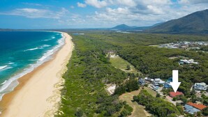 Vista aérea de uma área costeira com praia, floresta densa e um conjunto de casas com painéis solares.