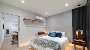 Une chambre moderne avec un grand lit, une table de chevet, un climatiseur fixé au mur et une armoire de couleur sombre.