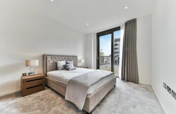 A modern bedroom with a large bed, a nightstand, a lamp, and a view of the city through a large window. - Londwell - Stunning Designer Penthouse, US Embassy (London)
