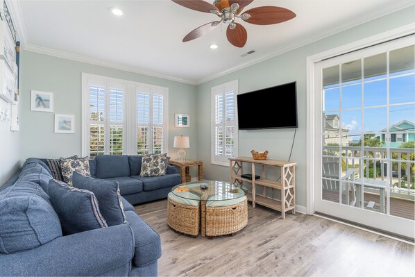 A living room with a blue sofa, wicker ottomans, a glass coffee table, a ceiling fan, a TV, and large windows with shutters.