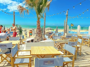A beachside restaurant with outdoor seating, palm trees, and a view of the beach.