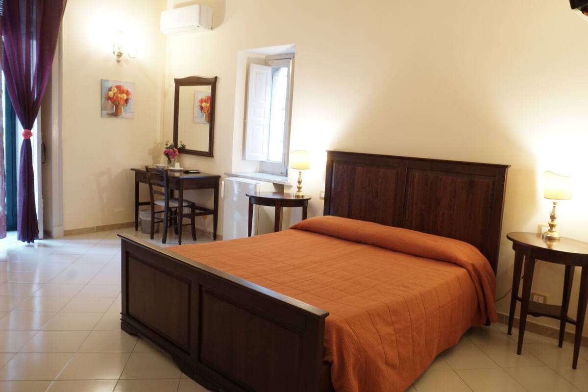 A bedroom with a wooden bed, a desk, a chair, a small table with a lamp, and a window with curtains.