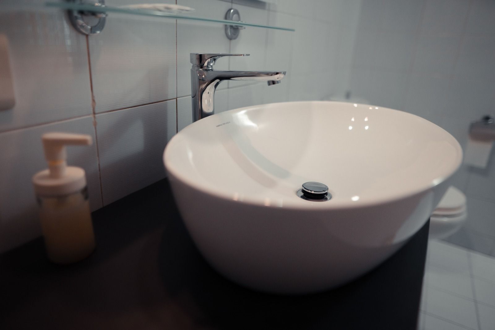A bathroom with a white sink, a soap dispenser, and a glass shelf above the sink.
