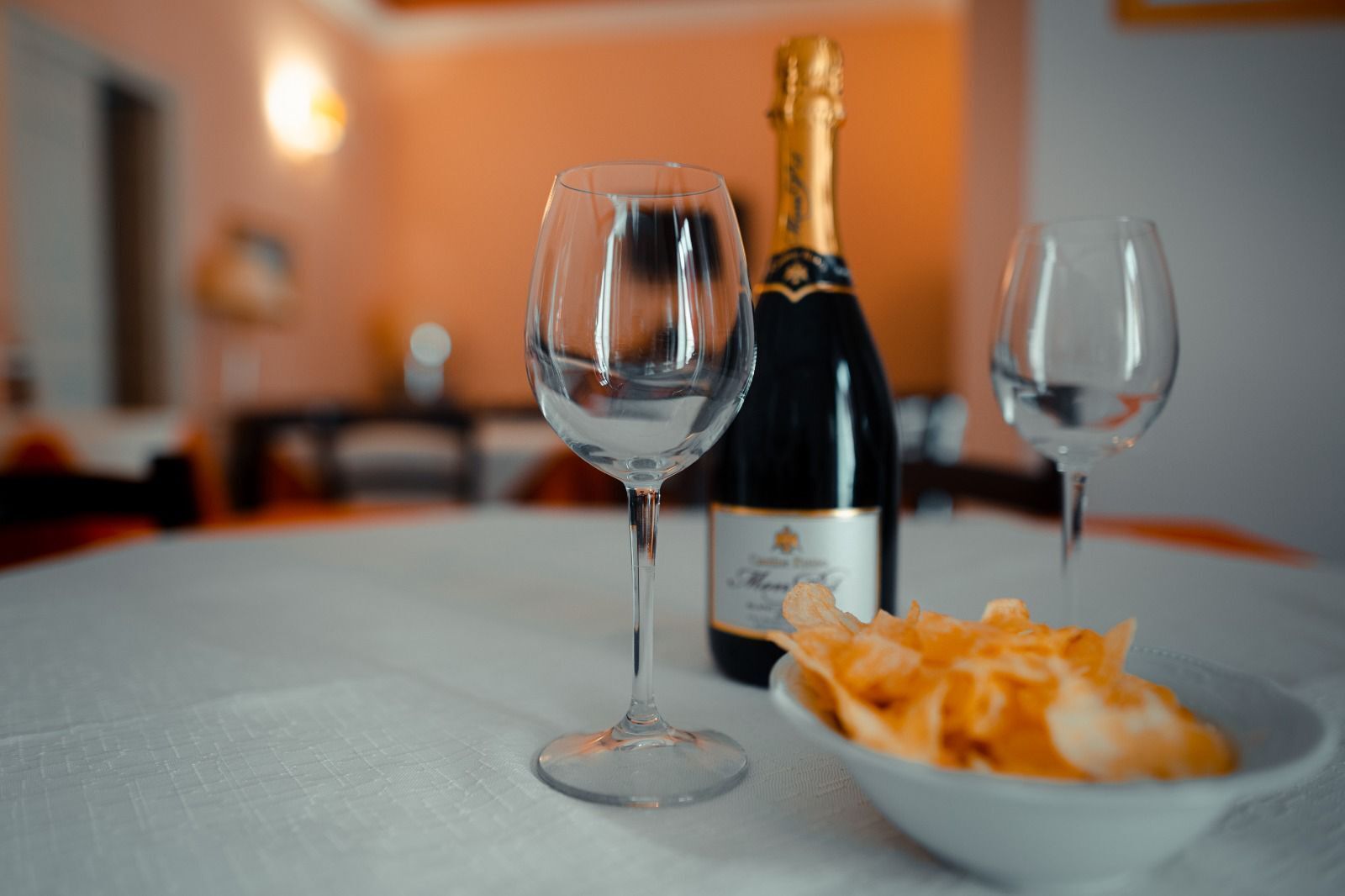 Una mesa con dos copas de vino vacías, una botella de champán y un bol de aperitivos.
