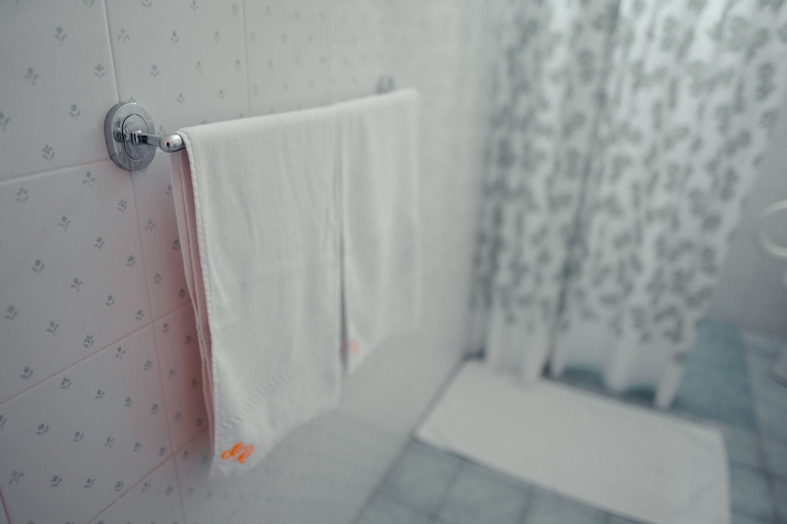 Un baño con pared de azulejos blancos, una toalla colgada en un perchero metálico y una cortina de ducha con estampado floral.
