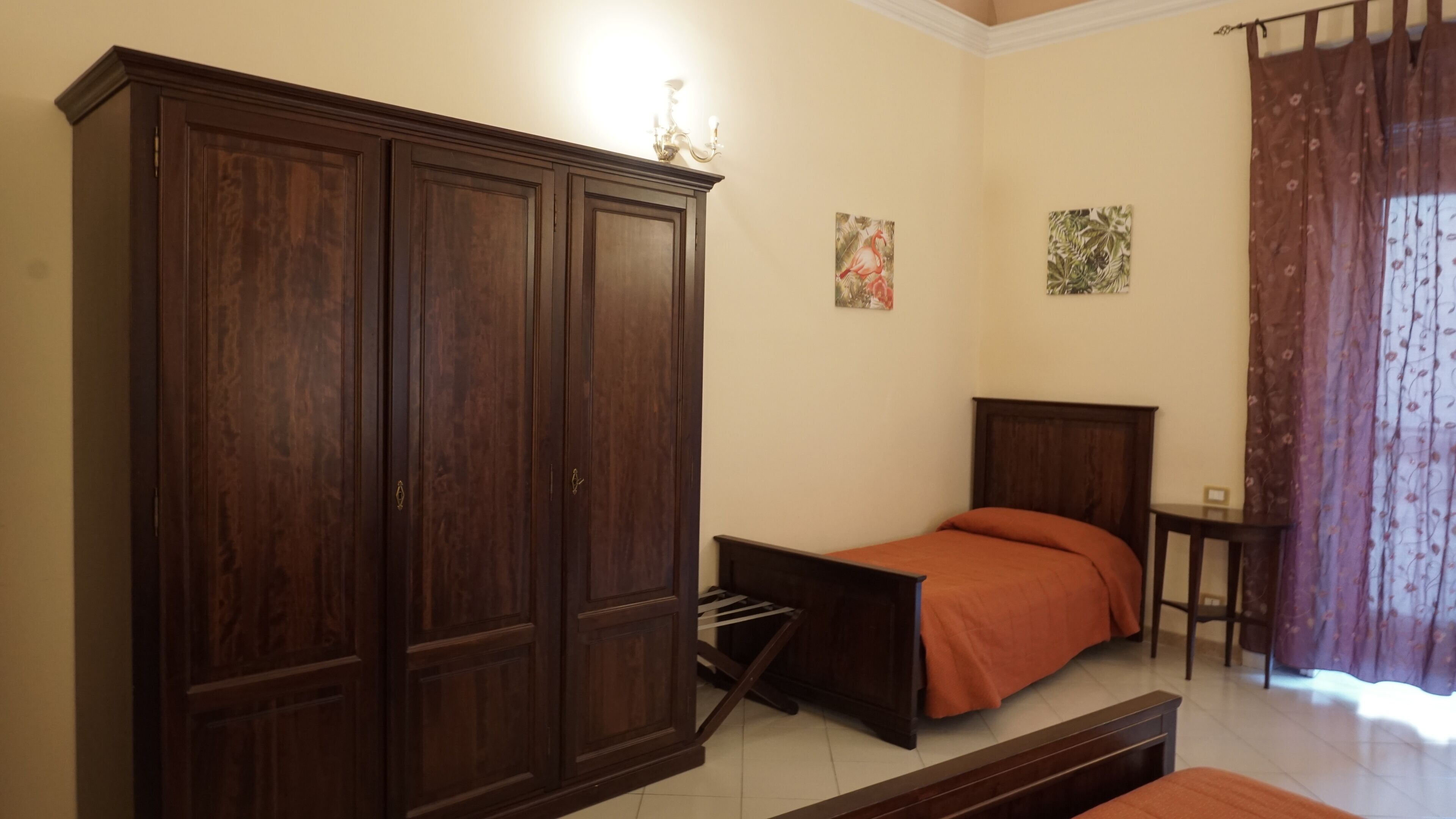 Un dormitorio con un armario de madera grande, una cama con colcha naranja, una mesita de noche pequeña y una mesita auxiliar.