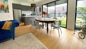 A modern kitchen with a central island, a dining area with a round table and patterned chairs, and a large sliding glass door leading to a backyard. - Capel Luxe (Capel Sound)