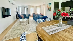 A modern living room with a flat-screen TV, a coffee table, sofas, and a dining area with a wooden table and chairs. - Capel Luxe (Capel Sound)