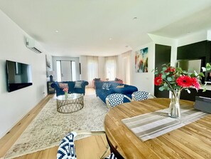 A modern living room with a flat-screen TV, a coffee table, sofas, and a dining area with a wooden table and chairs.