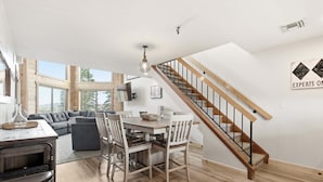 A modern kitchen with a built-in oven, a dining area with a wooden table and chairs, a staircase with wooden railings, and a large window offering a view of the outdoors.