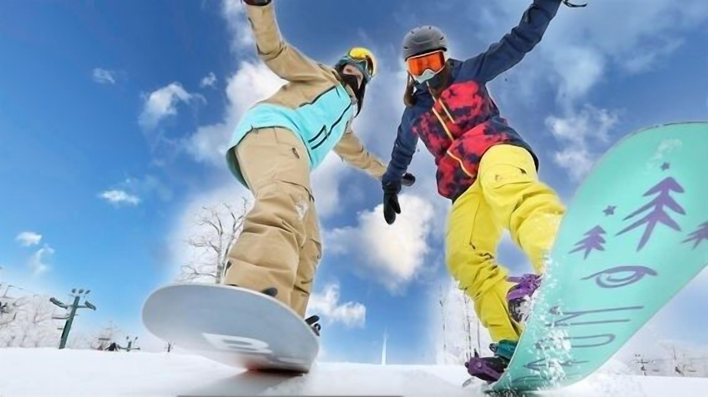 Zwei Snowboarder sind in Bewegung zu sehen vor blauem Himmel.