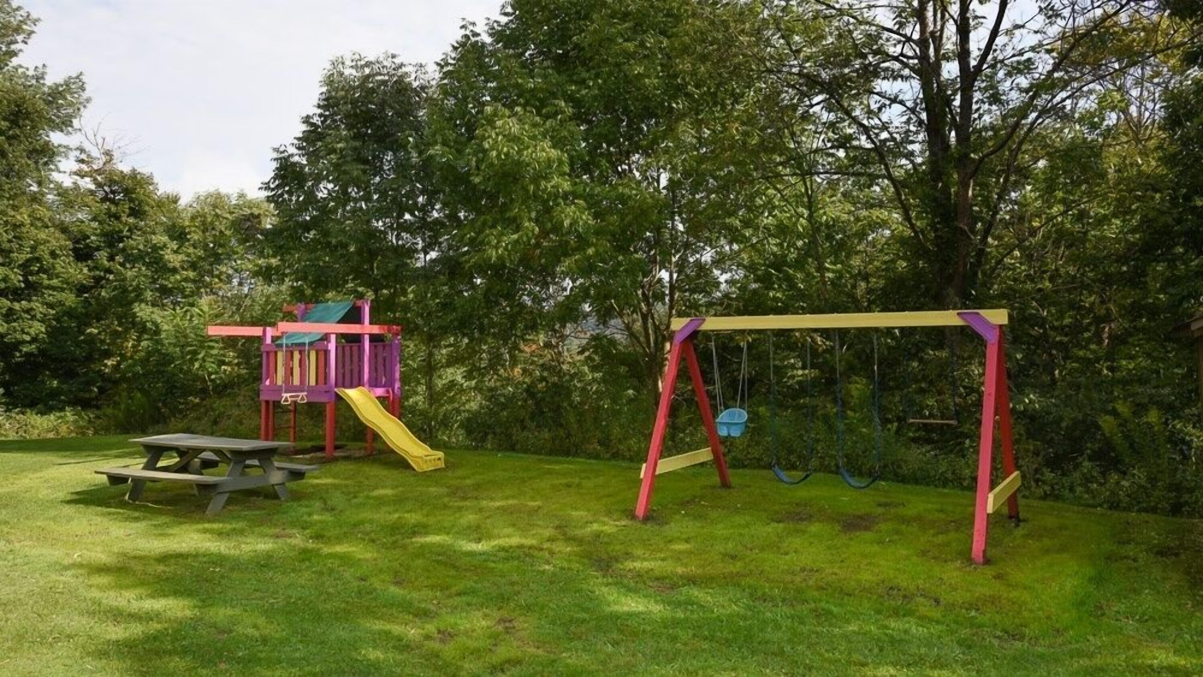 A playground with a slide and swings, surrounded by trees and grass.