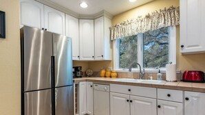 A kitchen with white cabinets, a stainless steel refrigerator, a sink, and a window with a view of trees. - Vineyard Cottage w/ Fire Pit | Chalk Hill Wine Country (Healdsburg)