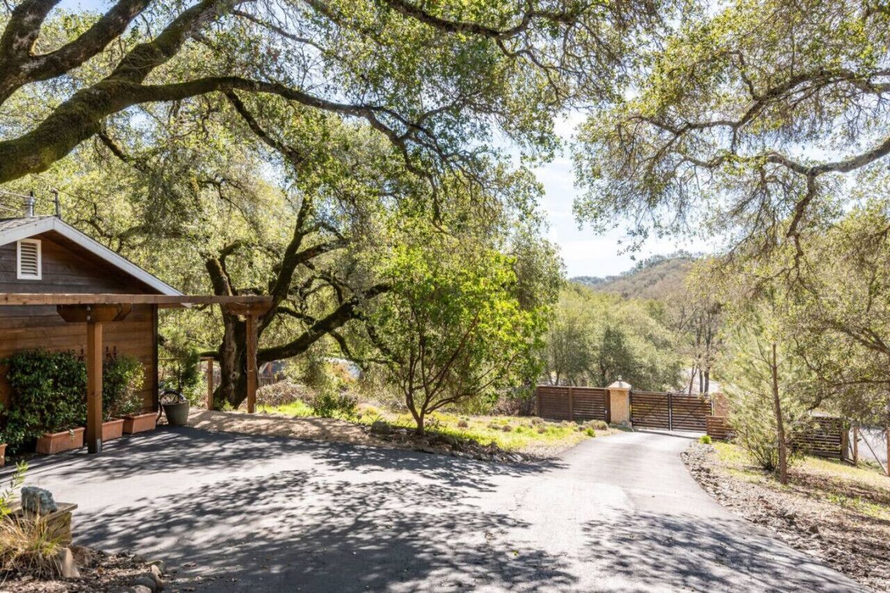 A rustic cabin with a covered porch, surrounded by trees and a dirt road leading to a gated entrance.