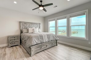 Un dormitorio con cama, mesitas de noche, ventilador de techo y ventanas amplias con vista al mar.