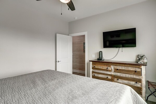 Un dormitorio con cama, cómoda de madera, televisor montado en la pared y puerta que da a otra habitación.