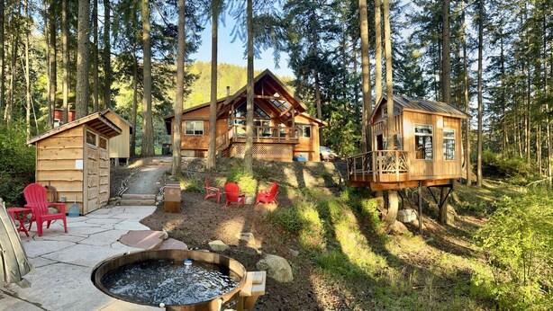 A cabin with a deck, a fire pit, and red chairs in a forest setting.