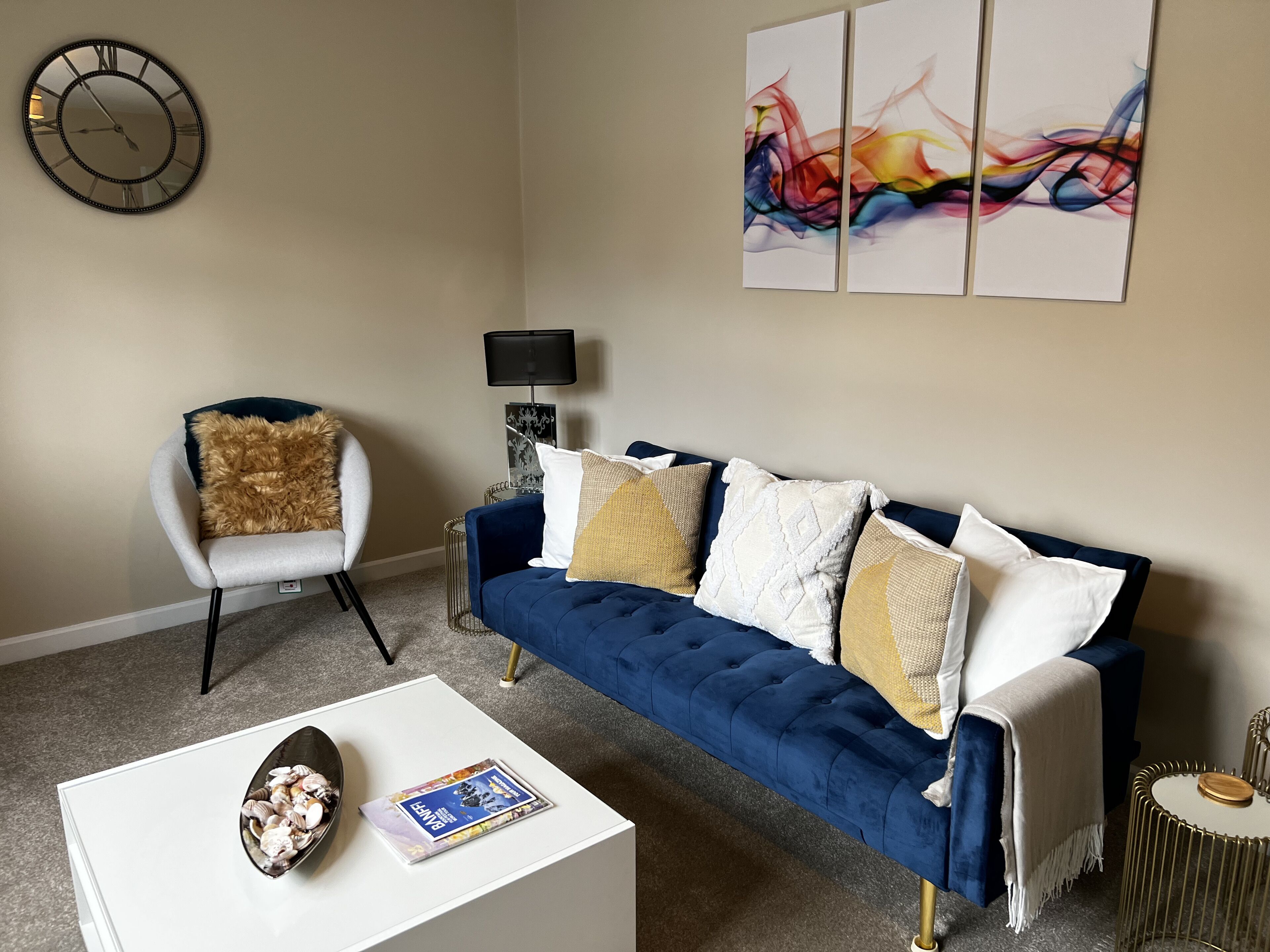 A living room with a blue sofa, a white armchair, a coffee table, a clock, and abstract art on the wall.