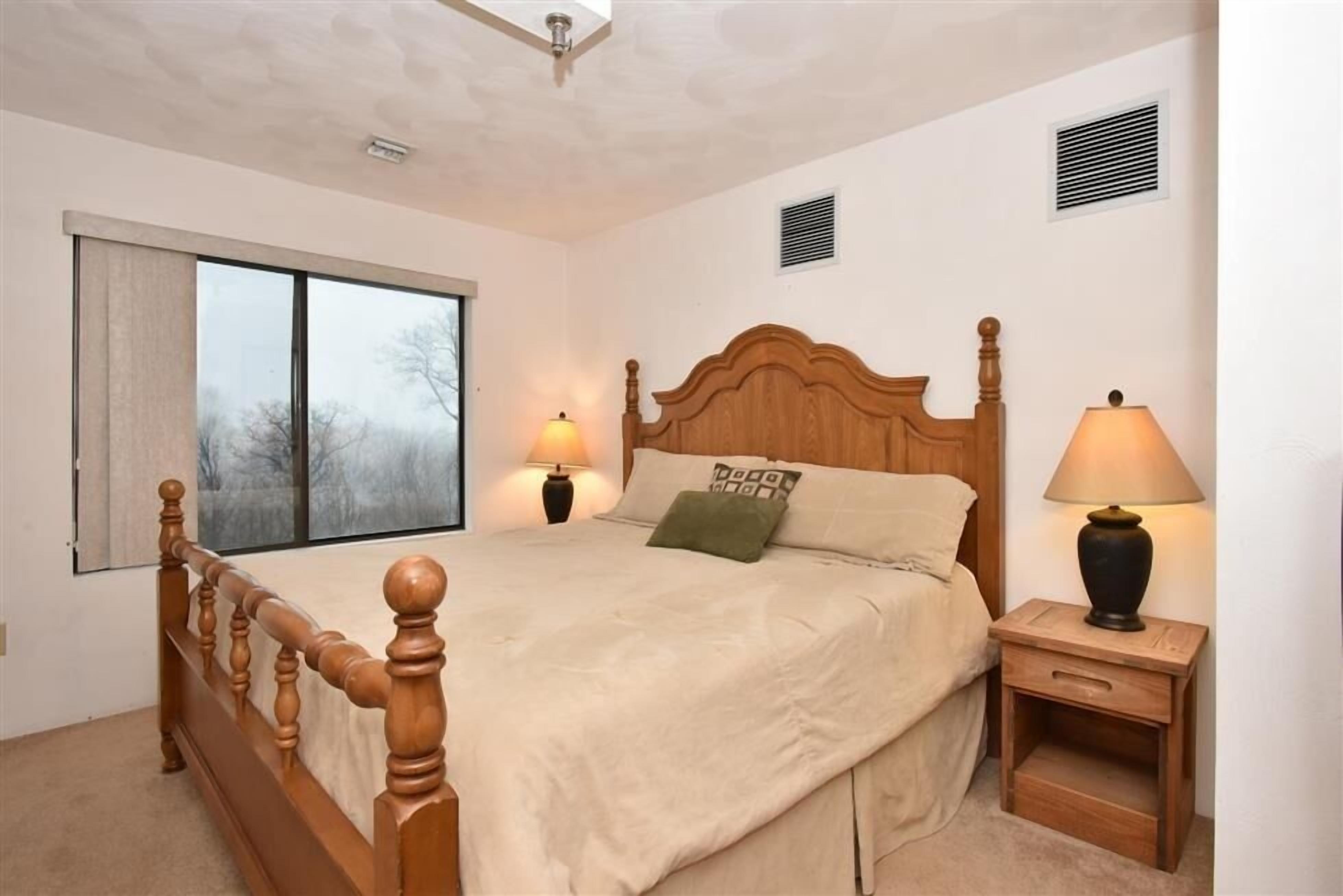 Une chambre à coucher avec un lit en bois, deux lampes de chevet, une table de nuit, une fenêtre avec des stores et un ventilateur de plafond.
