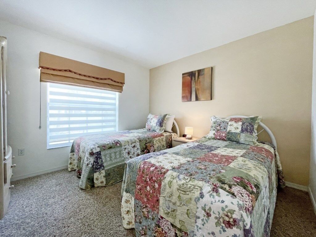 A bedroom with two beds, a window with blinds, a bedside table with a lamp, and a wall-mounted artwork.