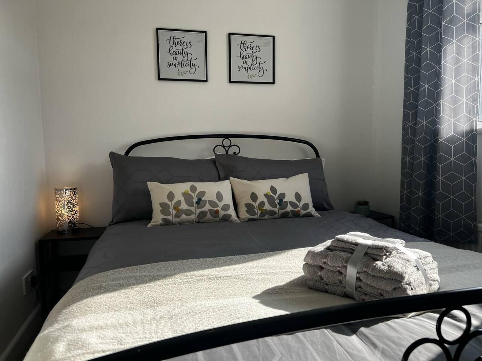 A neatly made bed with a grey bedspread and two pillows, a bedside lamp, and two framed inspirational quotes on the wall.