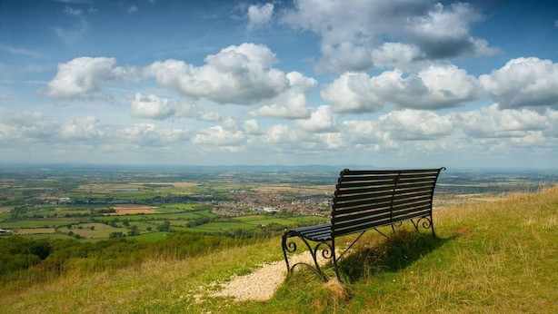 A bench on a grassy hill overlooking a valley with a town in the distance and a partly cloudy sky.