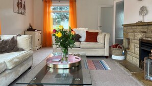 A living room with a glass coffee table, a sofa, a fireplace, and a view through a window with orange curtains.