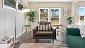 A living room with a leather armchair, a green sofa, a floor lamp, and a view of trees through large windows.