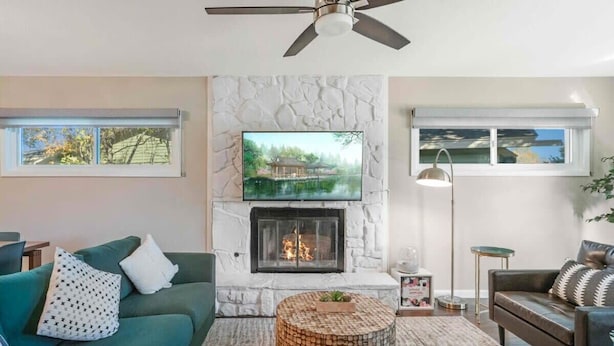 A living room with a stone fireplace, a ceiling fan, a television mounted above the fireplace, and a view of a house through the window.