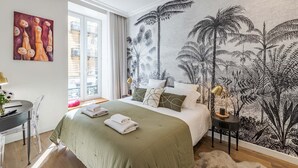 A bedroom with a bed, a desk, and a chair. There is a window with curtains, a wall with a tropical wallpaper, and a painting on the wall. - NEW-Hyper Centre-Cozy Chic-Renovated apartment with all comforts-AC-Balconnet-asc-Park (Nice)
