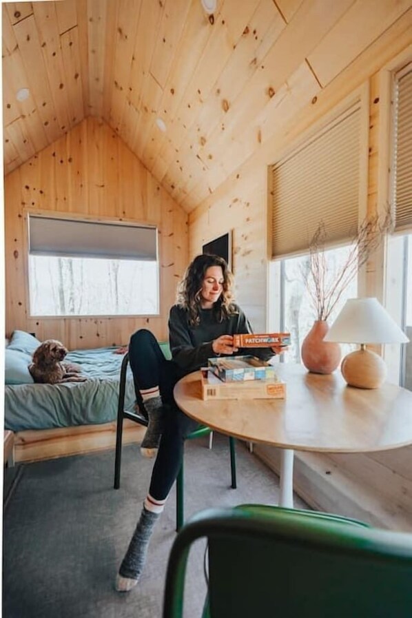 A cozy cabin interior with a wooden ceiling, a bed with a dog, a table with boxes, and a window with a view of trees. - Scandinavian Retreat, Mountain View, and 20 Minutes to Asheville! (Swannanoa)