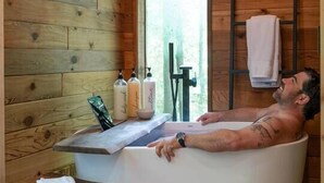 A man relaxing in a wooden cabin bathroom with a bathtub, a wooden bench, and a window. - The Twig, Outdoor Shower, Deck, and Cozy Chimney! (Sylva)