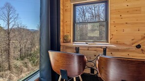 A wooden cabin with a window overlooking a forest. Inside, there is a table with two chairs and a small shelf with a book and a decorative item.