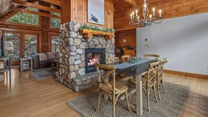 Een ruime woonkamer met een stenen openhaard, houten balken in het plafond en een eethoek met een tafel en stoelen.