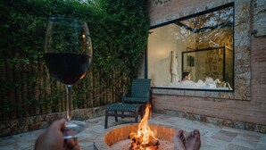 A person relaxing by a fire pit with a glass of wine, surrounded by a stone patio and a view of a brick wall and greenery. - Pousada LaPianta  (Camanducaia)