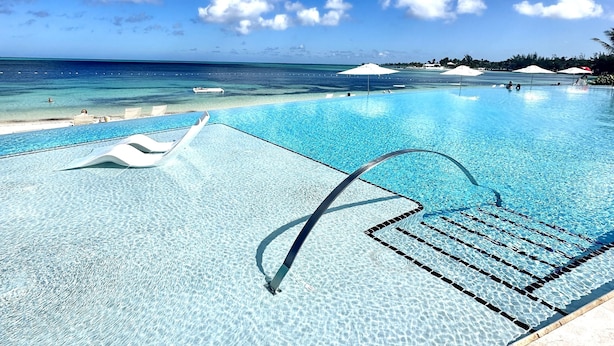 Piscina azul cristalina com plataforma de mergulho, cercada por uma praia e vista para o oceano.