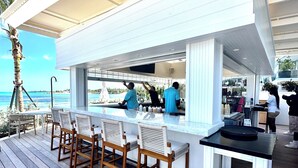 Área de bar ao ar livre coberta, com cadeiras e mesa de madeira, com vista para a praia.