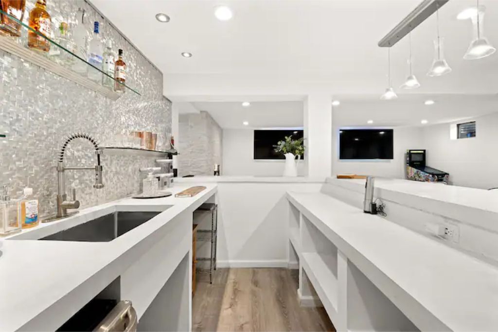 A modern kitchen with white countertops, a large sink, and a backsplash with a mosaic pattern. There is a window with a view of the night sky, and a television mounted on the wall.