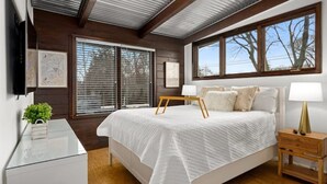 A bedroom with a large bed, wooden headboard, a bedside table with a lamp, and a window with blinds.